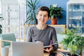 Smiling neutral gender middle-aged person making notes in her paper notebook, using laptop while working indoors in her workstation in an open space office. Online video call, IT HR, recruiting.