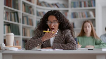 Young smart woman student in eyeglasses writing test and feeling tired, yawning and falling asleep on desk at classroom - Powered by Adobe