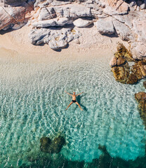 Fototapeta na wymiar Aerial photo of man lying on the water in STAR pose on rocky pebbly beach and sun tanning. Soft waves washing his body on lonely deserted Greek island in Ionian Sea . Exotic vacation concept