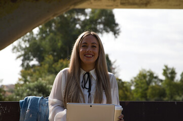 Portrait of Beautiful young blonde modern dressed woman sitting on a bench in the park with a laptop. The woman is holding an eco-friendly paper cup of coffee or infusion.
