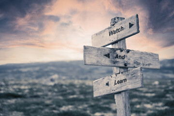 watch and learn text quote on wooden signpost outdoors in nature. Pink dramatic skies in the background.