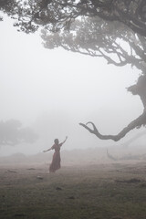 Barefoot woman in red dress dancing with eldar laurel tree looking like creature. Fanal forest, Madeira Island, Portugal, Europe.