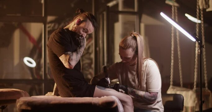 Female tattooist making painful tattoo on leg of bearded male client