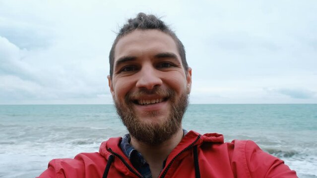 Lifestyle concept. Caucasian travel blogger stands on seashore and takes selfie video against waves on smartphone. Man in red jacket with beard and dreadlocks talking to camera. Portrait wide angle.