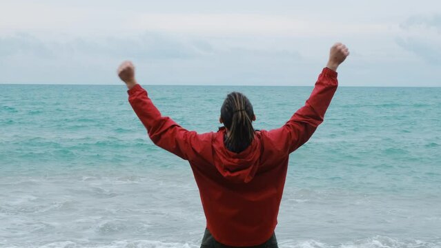 Lifestyle concept. Young Caucasian guy stands on seashore and rejoices with hands raised in different directions and up. Man in red jacket with dreadlocks enjoys views of coast in spring. Rear view.