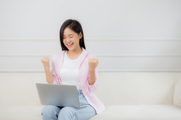 Happiness young asian woman sitting on sofa and working on laptop computer while successful and excitement at home, freelance female using notebook and achievement, business and lifestyles concept.