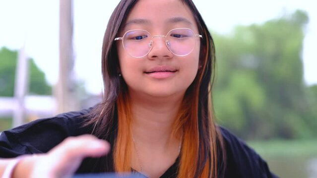 Asian girl with braces and glasses talking and playing mobile phone outdoors with smiling face.