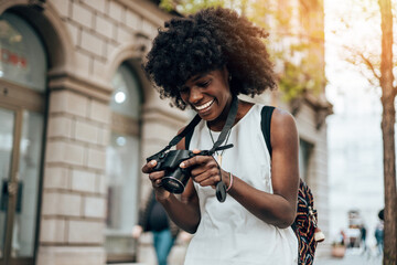 Young black female tourist enjoys walking through the streets of a beautiful European city. She is happy and using her photo camera to take fantastic architecture photographs. Bright sunny day.