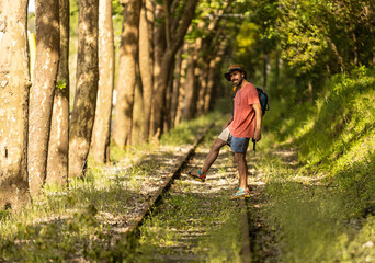 Fototapeta premium Backpacker man, walking along train tracks, observing Portugal's lush green forest in summertime. Wearing hat, pink t-shirt and short with sandals. Sunbeams.