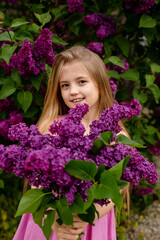 a beautiful happy smiling blonde girl with long hair in a pink dress with a purple bouquet of lilacs in her hands stands against the background of a lilac bush