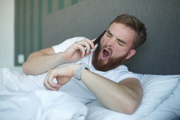 Young sleepy awaken man is just wake up with phone call, looking at his watches checking time and hurry up and yawn. Guy overslept job 