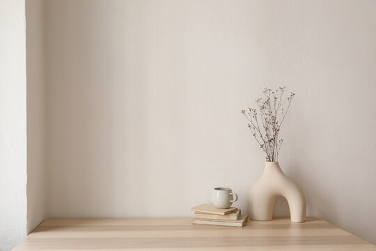 Elegant interior still life. Modern organic, geometric shaped vase with dry flowers, grass. Pile of old books, cup of coffee on wooden table. Home staging, minimal decor concept. Blank beige wall.