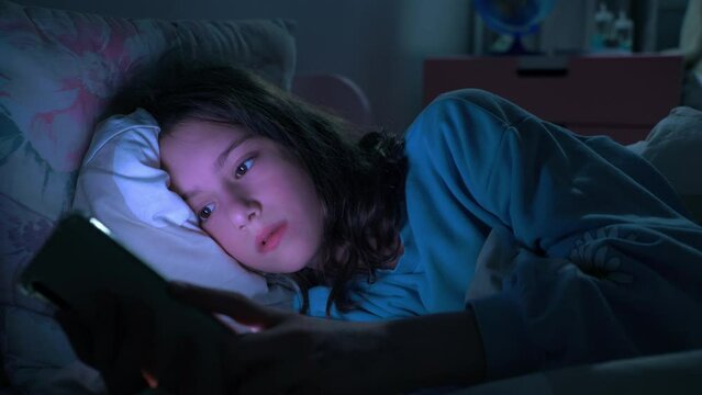 Teen girl scrolling social media texting on smartphone in bed in bedroom at night. Technology, internet, communication and people concept. Dependence on social networks in children. Not healthy sleep.
