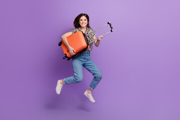 Fototapeta premium Full length portrait of overjoyed person jumping arms hold suitcase isolated on purple color background