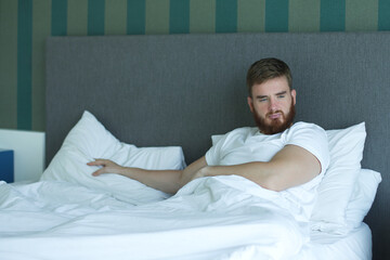 Young depressed unhappy bearded sleepy sad upset man lay in bed in bedroom at home in early morning