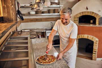 Smiling pizza owner, working like a pizza master, being in the kitchen, making delicious pizzas. - obrazy, fototapety, plakaty