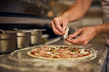 Male pizza maker putting mushrooms on the pizza dough, focus on the fresh made pizza dough.