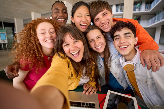 Selfie with mobile phone of multiracial cheerful group of students gathered in hall of university building. Happy young friends hugging laughing and posing for photo. People smiling on campus.