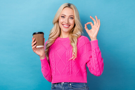 Photo of good mood pretty girl with wavy hairdo dressed pink sweater showing okey hold cup of latte isolated on blue color background