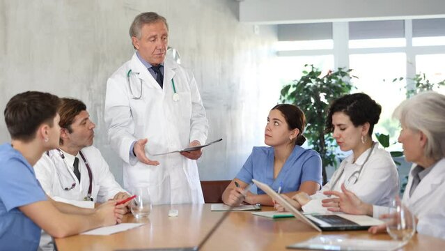 Focused experienced elderly head doctor conducting medical council with male and female colleagues of different ages sitting at table with papers and laptop in office. High quality 4k footage