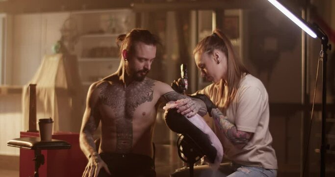 Focused female master making tattoo on arm of bearded male client drinking coffee