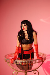 Pinup girl standing near big glass. Black wig, black underwear, stockings and red gloves, red and blue light, red lipstick. Beautiful posch woman sitting near huge plastic goblet.