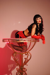 Pinup girl standing near big glass. Black wig, black underwear, stockings and red gloves, red and blue light, red lipstick. Beautiful posch woman sitting near huge plastic goblet.