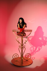 Pinup girl standing near big glass. Black wig, black underwear, stockings and red gloves, red and blue light, red lipstick. Beautiful posch woman sitting near huge plastic goblet.