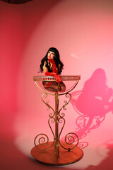 Pinup girl standing near big glass. Black wig, black underwear, stockings and red gloves, red and blue light, red lipstick. Beautiful posch woman sitting near huge plastic goblet.