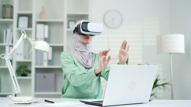 Muslim business woman employee worker entrepreneur wearing virtual reality VR headset glasses using metaverse technology to visual design project gestures with hands at desk in office workplace Indoor