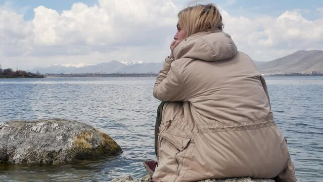An adult young blonde is sitting on large stones on the seashore and looks into the distance at the water and high snowy mountains. Woman in a jacket sitting on a smooth round stone on the shore