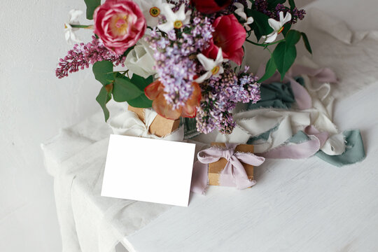 Greeting card mock up and lilac, tulips, daffodils flowers with present and pastel ribbons in modern rustic room. Happy Mothers day. Space for text. Stylish bouquet with gift box on wooden table