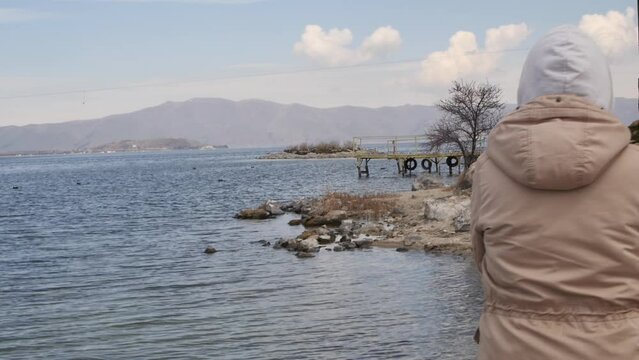 A young woman walks along the seashore wearing a warm jacket and hood. An adult woman walks alone on a rocky seashore and seagulls fly over the water