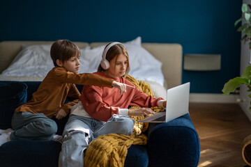 Teen girl sister in headphones and brother and sister watching movie together on laptop sitting on couch at home. Boy pointing something on screen. Leisure, digital entertainment, recreation concept.