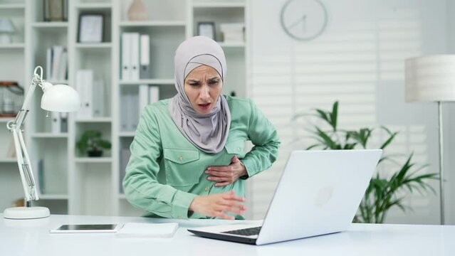 Muslim female has a heart attack pain, tension Sick employee feeling heartache while working on laptop at workplace at desk in office Sad woman in hijab keeps her hand on her chest heartattack indoor