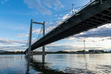 A cable bridge spans across a river against a cloudy blue sky at sunset.