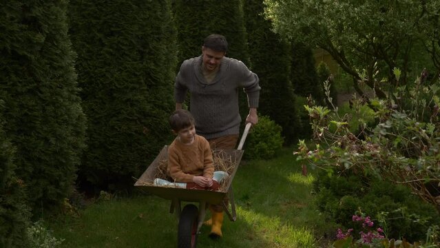 father rides son in a wheelbarrow with hay in the garden in spring time, having fun together