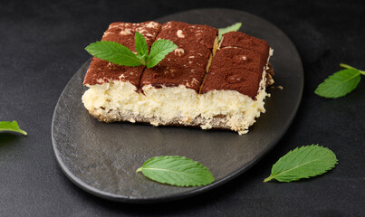 A piece of tiramisu sprinkled with cocoa, on top of a sprig of fresh mint on a black background