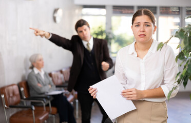 Upset young woman in business clothes with documents in her hands being criticized in office
