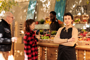 Young confident woman wearing apron running successful farmers market stand, selling fresh organic produce directly to customers. Female greengrocer standing with crossed arms and looking at camera