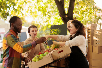 Vendor offering samples to customers while selling home-grown fruits and vegetables at local farmers market. Young multiracial family couple tasting natural organic produce while visiting food fair - obrazy, fototapety, plakaty