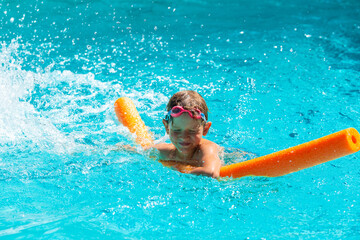 Oudoor summer activity. Concept of fun, health, vacation. Happy smiling boy five years old in swimming goggles laught and closes himself from splashing water in the pool with noodle on hot summer day.