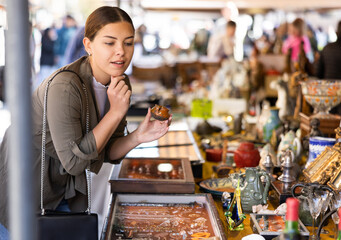 Satisfied interested young woman choosing interesting antique things at traditional flea market