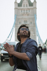 Close up handsome stylish guy in London is opposite of London Bridge, man standing on Tower Bridge. He wears a denim jacket, sunglasses and he is looking away from camera.