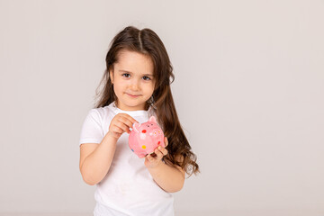 Positive girl in casual outfit laughing and saving money in piggy bank against white background