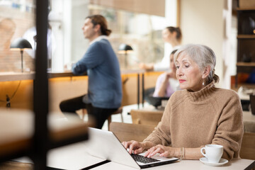 Positive European old woman drinking and enjoying coffee while working on laptop in cafe