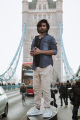 Handsome stylish guy in London is opposite of London Bridge, man standing on Tower Bridge. He wears a denim jacket, sunglasses and he is looking away from camera.