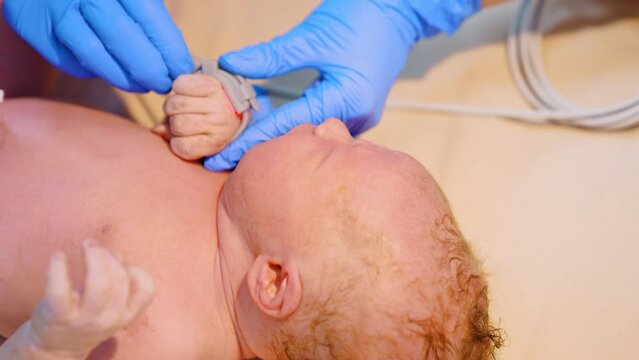 Caucasian newborn baby cries while medic attaches medical band on its hand. Close up.