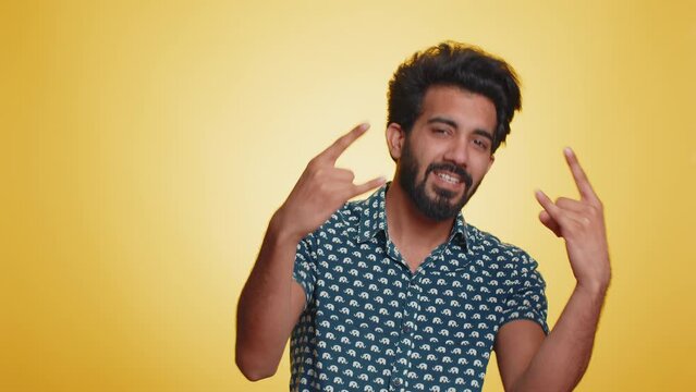 Overjoyed young indian man showing rock n roll gesture by hands, cool sign, shouting yeah with crazy expression, dancing, emotionally rejoicing in success. Hindu guy isolated on yellow background