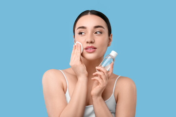 Beautiful woman removing makeup with cotton pad on light blue background